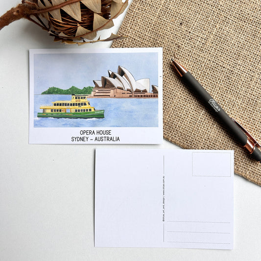 Postcard with Sydney Opera House illustration on a textured surface with a pen and decorative plant.