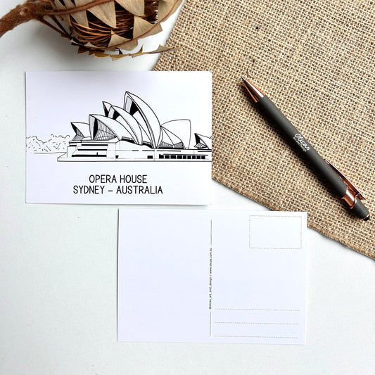 Postcard with Sydney Opera House illustration, pen, and decorative plant on textured surface